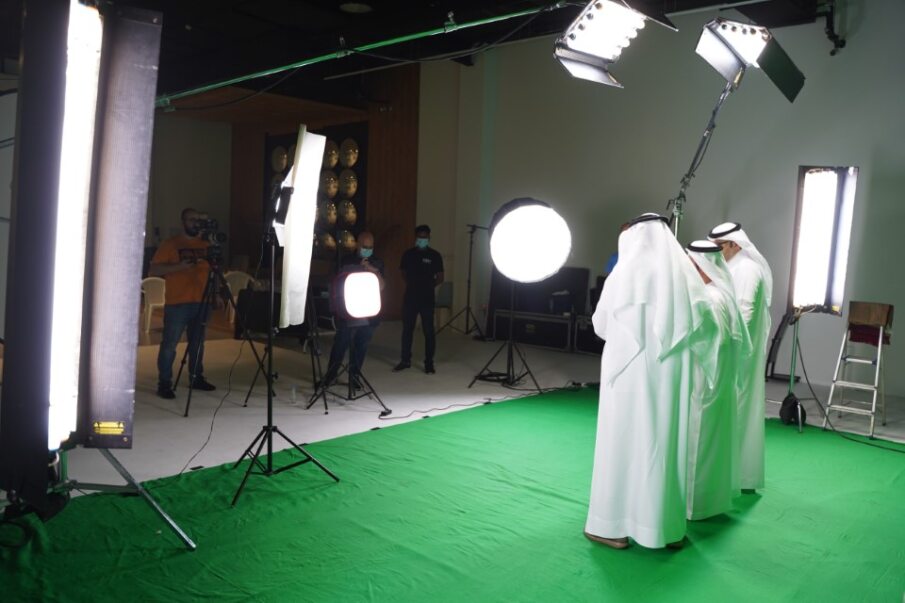 two men in traditional white attire stand in front of a green screen, and a camera setup highlights a media production