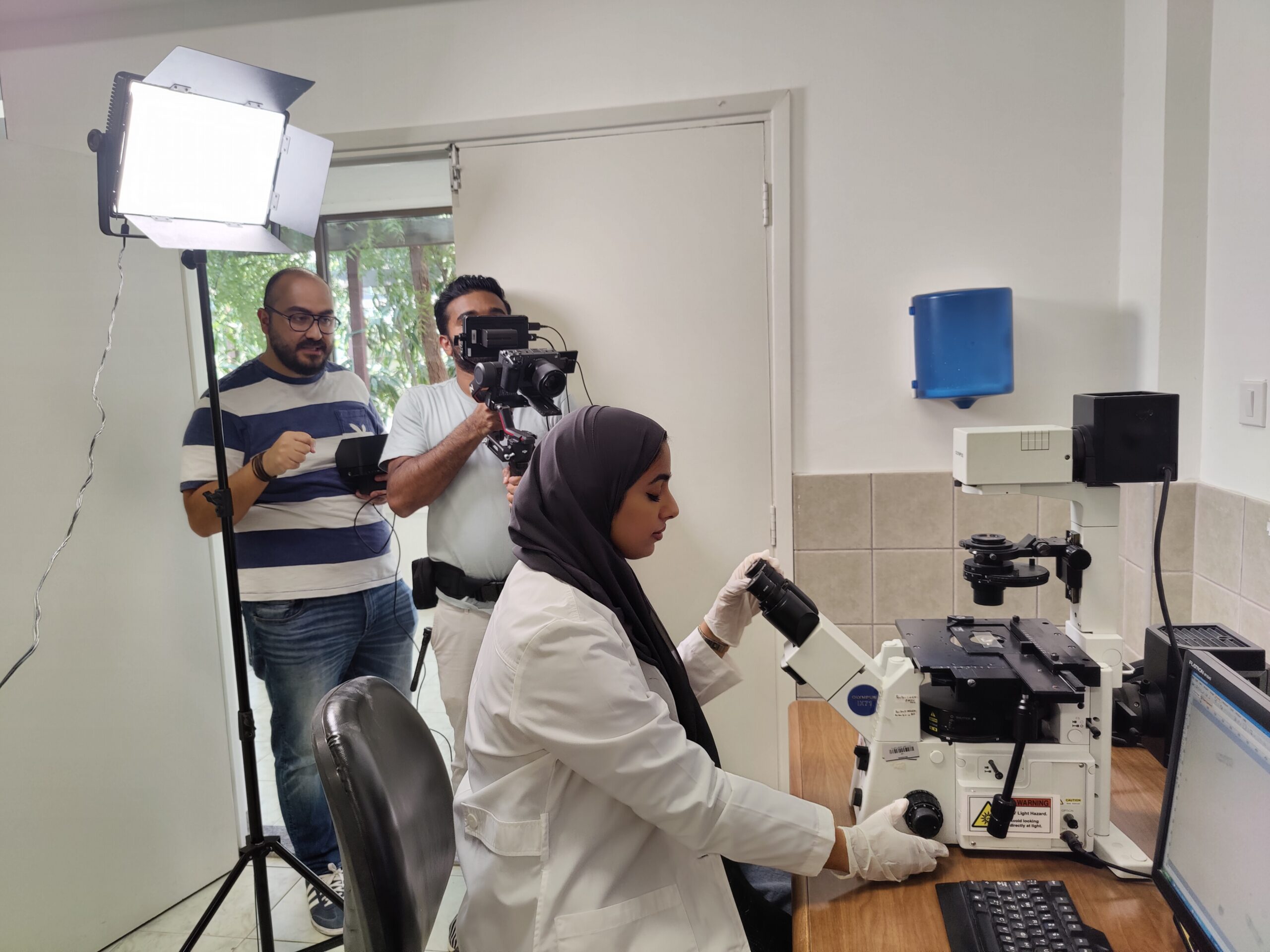 woman sits on stool in professional photo shoot setup. Setting appears office with large windows and cityscape view outside.