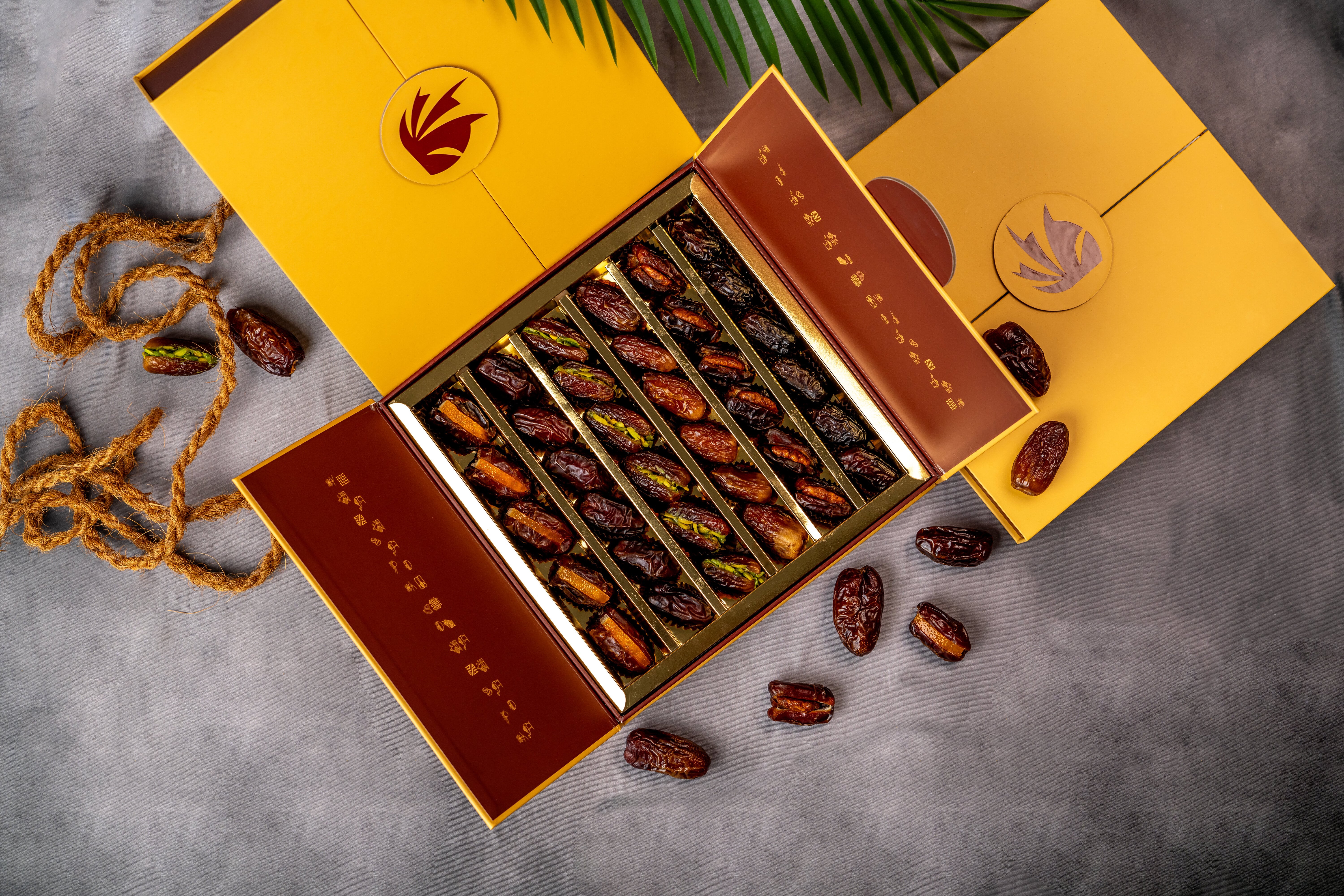 A fancy open box of assorted dates with some spilled onto a grey surface, alongside another closed similar box.