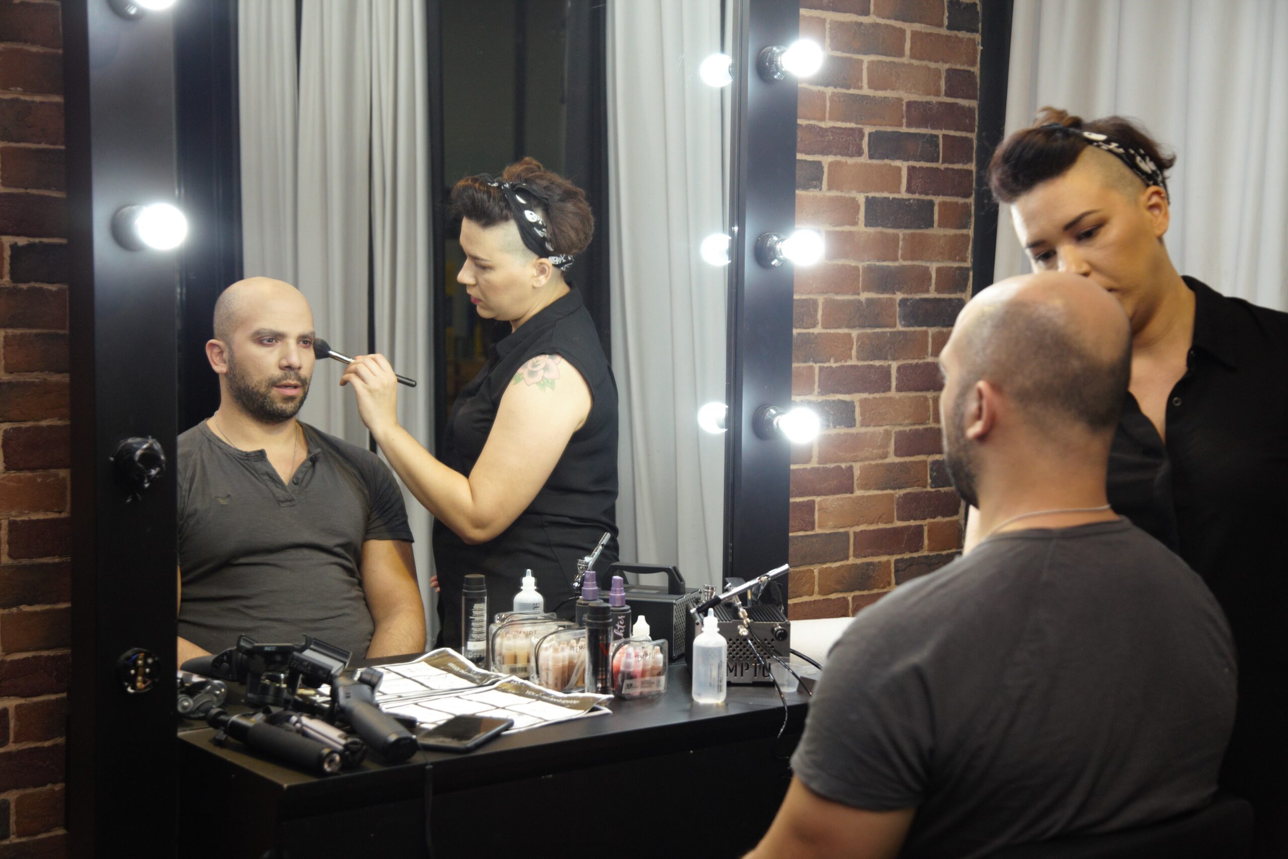 A woman sits in a chair reading a magazine in front of a backdrop, while a photographer adjusts equipment nearby.